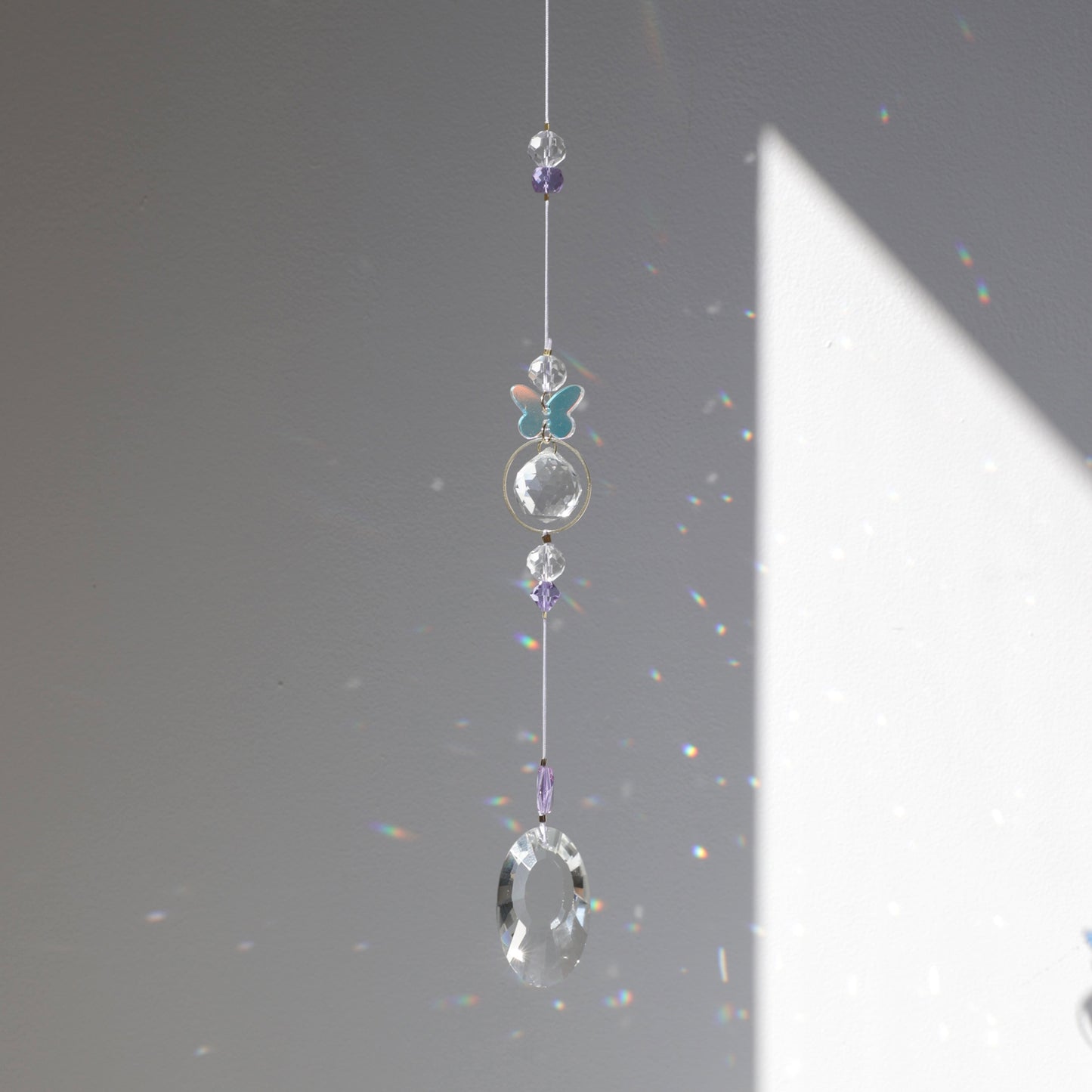 Suncatcher Purple Aura de la marque Rayonne avec papillon holographique,suspendu près d’une fenêtre, projetant des reflets arc-en-ciel à la lumière du soleil.
