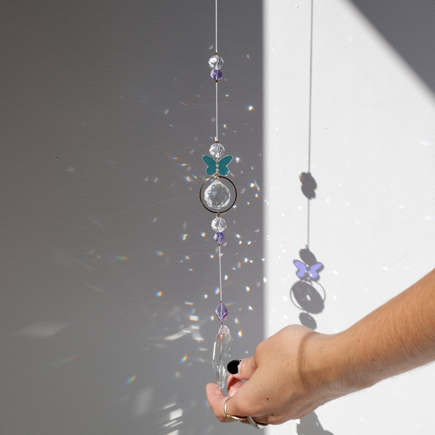 Suncatcher Rayonne® avec papillon suspendu près d’une fenêtre, projetant des reflets arc-en-ciel.