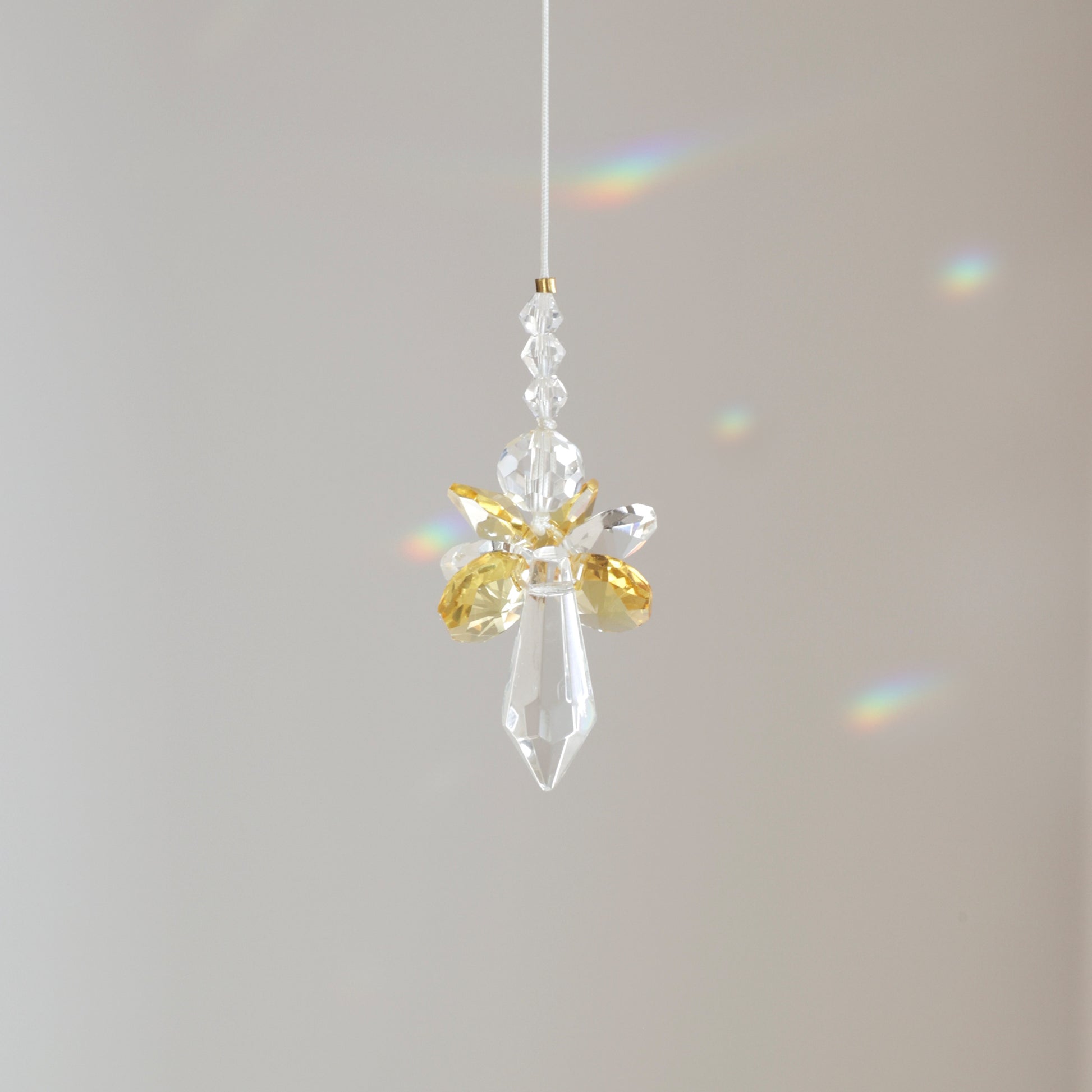 Suncatcher ANGE, symbole de joie et d’énergie, suspendu à une fenêtre ensoleillée et diffusant des reflets arc-en-ciel. Cadeau de naissance