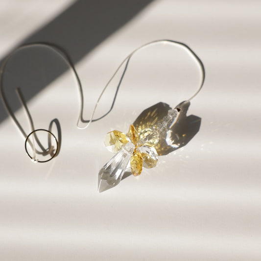 Gros plan sur le Suncatcher ANGE en cristal, captant la lumière du soleil et projetant des prismes lumineux colorés. Cordon ajustable pour une installation facile.