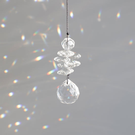 Mini Suncatcher Black Aura de la marque Rayonne® suspendu près d’une fenêtre, reflétant des couleurs arc-en-ciel dans une pièce lumineuse.