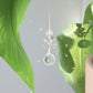 Mini Suncatcher Pink Aura suspendu à une plante près d’une fenêtre, projetant des reflets arc-en-ciel dans la pièce.