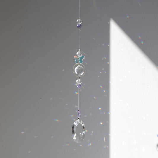 Suncatcher Purple Aura de la marque Rayonne avec papillon holographique,suspendu près d’une fenêtre, projetant des reflets arc-en-ciel à la lumière du soleil.