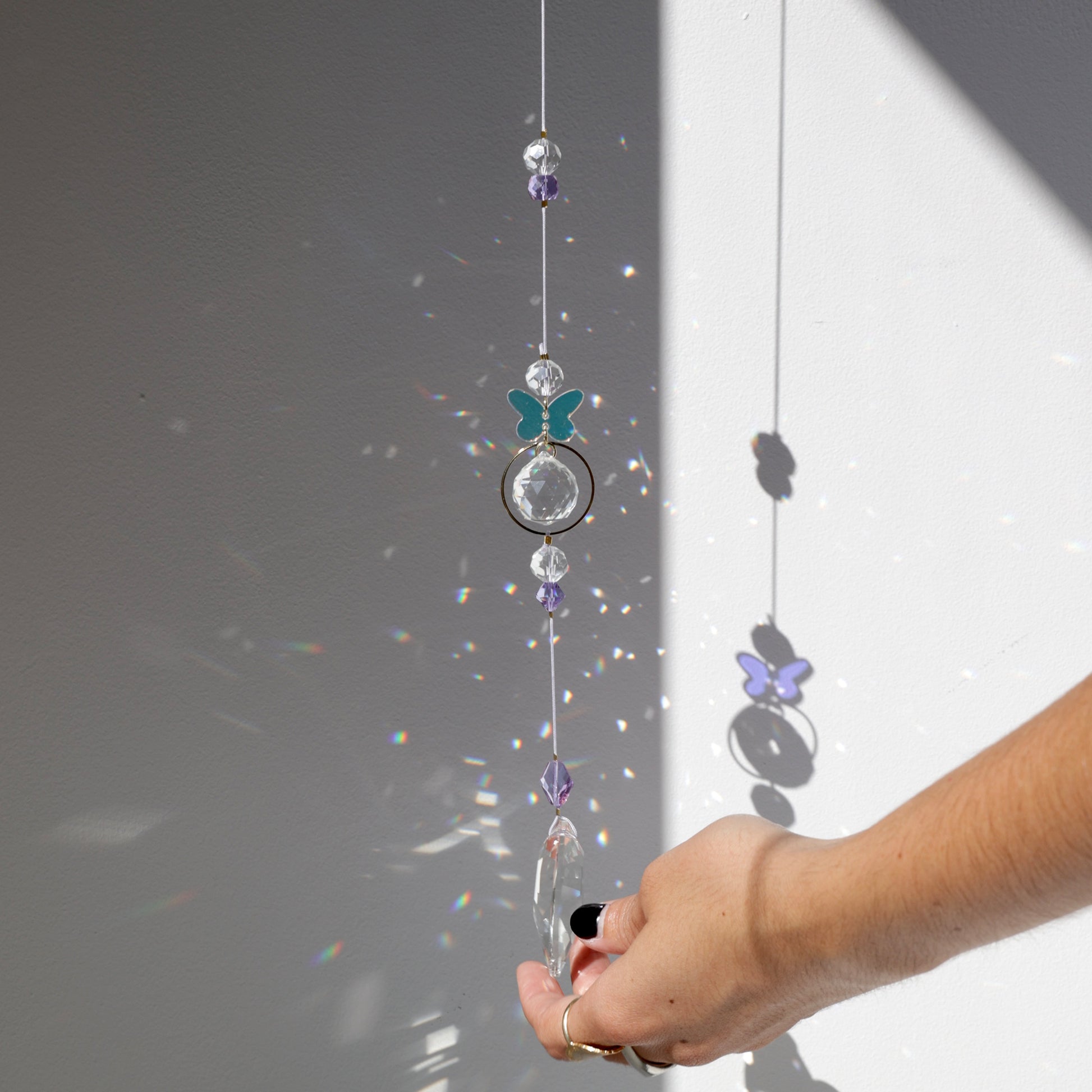 Suncatcher Rayonne® avec papillon suspendu près d’une fenêtre, projetant des reflets arc-en-ciel.