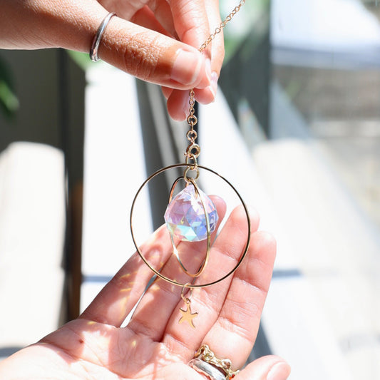 Suncatcher SUNSET suspendu devant une fenêtre ensoleillée, projetant des reflets arc-en-ciel sur le mur. 